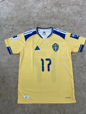 Svensk landslagströja Gyökeres 17 - Gul svensk landslagströja från Adidas med blå detaljer, nummer 17 och namnet Gyökeres på ryggen. Tröjan har blått landslagsemblem på bröstet, blå Adidas-logga och Climacool-material för bra ventilation. Korta ärmar med blå ränder och patchar på båda ärmarna.