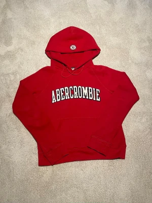 Abercrombie & fitch Vintage - Det står M men passar Xs-S. Säljer nu denna supersnygga och klassiska hoodie från Abercrombie & Fitch i en riktigt härlig röd färg! Tröjan har den ikoniska broderade loggan över bröstet i vitt och marinblått, samt en liten logga på huvan. Den har en perfekt vintage/Y2K-vibe och är gjord i ett rejält, mjukt material som håller värmen bra. Raglanärmarna ger den en snygg passform. • Skick: Mycket fint begagnat skick, väl omhändertagen! • Färg: Klarröd med vita detaljer.