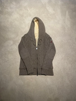 Stickad hoodie med fuskpälsfoder - Mysig grå, brun stickad hoodie med dragkedja och tjockt fuskpälsfoder i beige. Har kabelstickat mönster, ribbade muddar och en broderad detalj på ärmen. Perfekt för kalla dagar och riktigt skön att ha på sig.