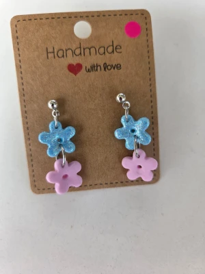 Handgjorda blommiga örhängen - Supersöta handgjorda örhängen med två retroblommor i rad – en glittrig blå och en matt rosa. Blommorna hänger från små silverfärgade kulstift av rostfritt stål. Jättefina till sommaren! Gjorda för hand av mig i polymerlera. 