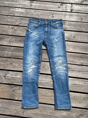 Blåa nudie jeans lean dean - Säljer ett par blå jeans med raka ben och slitningar på både fram- och baksida. Jeansen har klassisk femficksmodell och en ljus tvätt med tydliga slitdetaljer för en trendig look. Perfekta för dig som gillar en avslappnad och cool stil. Modellen är lean Dean