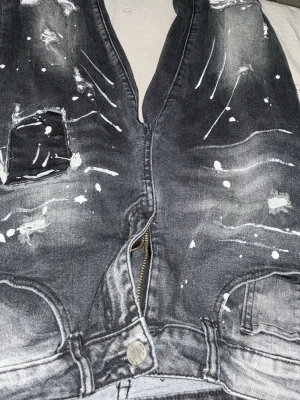 Svarta jeans med vit färg och slitningar - Säljer ett par svarta jeans med coola vita färgstänk och slitningar framtill. Jeansen har patchdetalj på ena benet och klassisk femficksdesign. Materialet är denim och passformen är rak. Perfekta för dig som gillar en edgy streetstil.