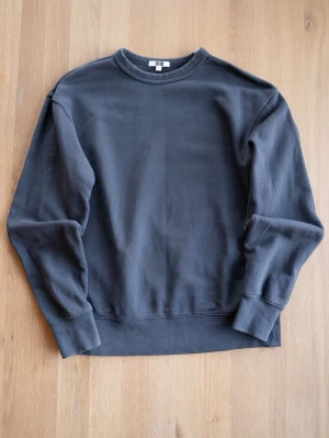 Mörkgrå sweatshirt från Uniqlo, size S/M - En stilren mörkgrå sweatshirt från Uniqlo U med rund halsringning och långa ärmar. Tröjan har ribbade muddar vid ärmslut och nederkant. Tillverkad i mjuk bomull för en bekväm känsla och enkel att matcha med olika outfits.