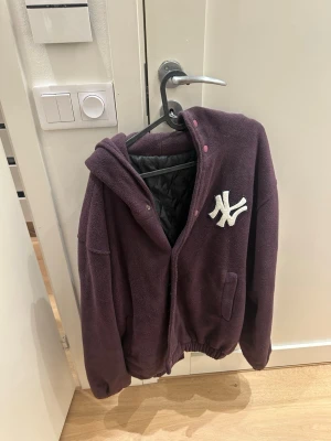 Lila teddyjacka med NY-logga - Mysig lila teddyjacka med huva och broderad vit NY-logga på bröstet. Jackan har dragkedja, tryckknappar, två fickor och quiltat svart innerfoder. Perfekt för dig som vill ha en sportig och trendig look.