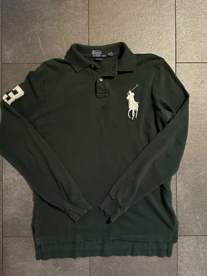 Mörkgrön långärmad piké från Polo Ralph Lauren - Klassisk mörkgrön långärmad pikétröja från Polo Ralph Lauren med stor vit broderad logga på bröstet och siffran 3 på ärmen. Skjortan har krage, knappar vid halsen och är tillverkad i bomull. Perfekt för en stilren och sportig look.