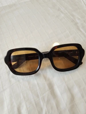 Svarta amber fyrkantiga solglasögon - Snygga solglasögon från Ace & Tate med bred, svart båge och fyrkantig form. Glasen är bruna och ger en cool retrovibe. Perfekta för att lyfta din stil under soliga dagar.