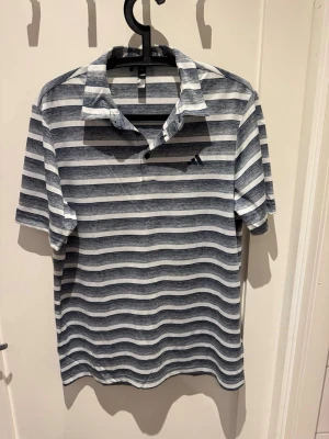 Pike adidas - Snygg randig pikétröja från Adidas i blått och vitt. Skjortan har korta ärmar, klassisk krage och knappar vid halsen. Perfekt för en avslappnad och sportig look. Tillverkad i mjukt och bekvämt material. Används som en Golfpike men funkar som vanlig också. Helt ny. 