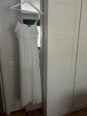 Vit långklänning från BikBok - Säljer en elegant vit långklänning från BikBok med smala axelband och volangdetaljer upptill. Klänningen har en luftig passform och är perfekt för sommarens alla tillfällen. Tillverkad i ett lätt och mjukt material som ger en skön känsla. Perfekt till skolavslutningen 