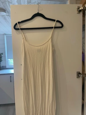 Lång vit klänning med tunna band - Säljer en enkel och stilren lång vit klänning med tunna axelband och rak passform. Klänningen har en minimalistisk look och är perfekt för varma dagar. Materialet känns lätt och svalt, troligen bomull eller polyester.