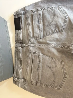Gråa jeans från ITB Valérie - Snygga gråa jeans från ITB Valérie med klassisk femficksmodell och raka ben. Jeansen har en ljus tvätt och detaljerade sömmar på bakfickorna. Tillverkade i mjukt denimtyg med knappgylf och bälteshällor. Perfekta för en avslappnad och trendig look.