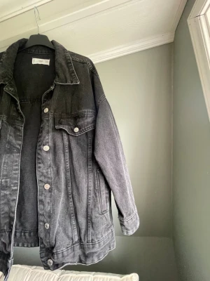 Svart oversized jeansjacka från Monki - Säljer en svart oversized jeansjacka från Monki med klassisk krage, silverfärgade knappar och två bröstfickor med lock. Jackan har en avslappnad passform och är tillverkad i kraftigt denimtyg. Perfekt för en cool och avslappnad stil.