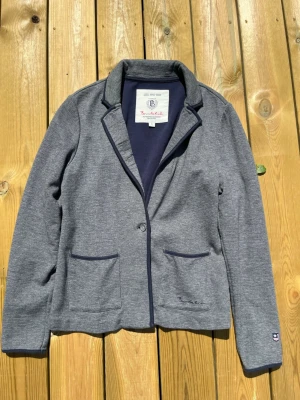 Grå kavaj från Bondelid, storlek S - Snygg grå kavaj från Bondelid i mjukt sweatshirtmaterial. Klassisk modell med två framfickor, enkel knappstängning och mörkblå kantband. Perfekt för en avslappnad men stilren look. Märkeslogga på ärmen och broderad detalj vid fickan.