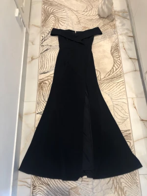 Marinblå offshoulder långklänning - Elegant marinblå långklänning med offshoulder-design och slits framtill. Klänningen har en figurnära passform upptill och faller snyggt mot golvet. Perfekt för dig som vill ha en stilren och sofistikerad look. Bilderna från hemsidan har inte samma färg på klänningen, då min är marinblå och den från hemsidan är svart🥰Använd endast en gång😊