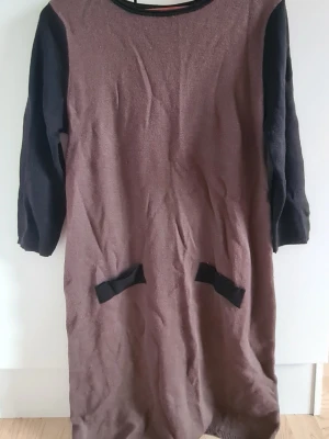 Brun stickad klänning med svarta ärmar - Mysig brun stickad klänning med svarta långärmar och svarta detaljer vid fickorna. Rund halsringning och rak passform, perfekt för en avslappnad stil. Klänningen har två dekorativa fickor framtill och är i ett mjukt material.