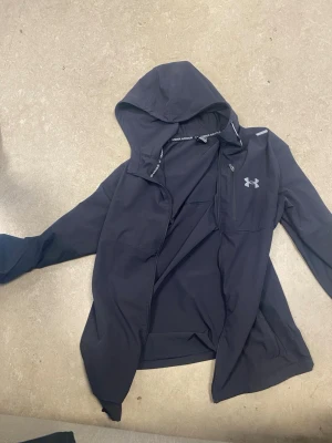 Svart vindjacka från Under Armour - Svart vindjacka från Under Armour med huva och dragkedja. Jackan har en diskret logga på bröstet och är tillverkad i ett lätt, vattenavvisande material. Perfekt för blåsiga dagar och enkel att matcha med olika outfits.