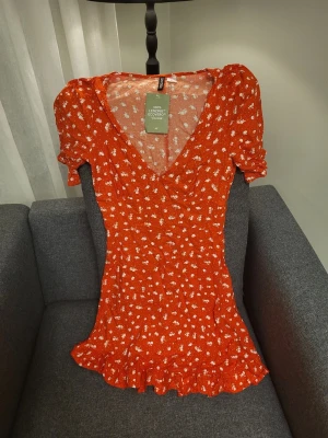 H&m Divided orange sommar klänning, storlek XS - Supersöt orange kortklänning från H&M med vitt blommönster, v-ringad hals och volangkant nertill. Klänningen har korta puffärmar och är tillverkad i 100% LENZING™ ECOVERO™ viskos. Perfekt för sommarens alla tillfällen!
