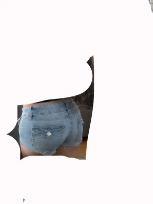 Ljusblå jeansshorts med fickor i baksidan  - Snygga ljusblå jeansshorts med låg midja och råa kanter. Klassisk femficksmodell med knapp och dragkedja framtill samt bakfickor med knappdetalj. Perfekta för varma sommardagar och enkel att matcha med olika toppar.