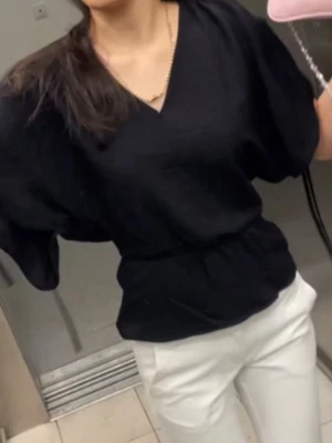 Svart blus med v-ringning från H&M - Svart blus med v-ringning och vida, korta ärmar med knytband i sidorna. Modellen har markerad midja och lätt volang nedtill. Perfekt för en trendig och avslappnad look.