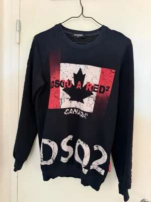 Svart sweatshirt från Dsquared2 - Cool svart sweatshirt från Dsquared2 med stort tryck av Kanadas flagga och texten DSQ2 på bröstet och ärmarna. Tröjan har rund halsringning och långa ärmar. Materialet känns mjukt och bekvämt, perfekt för en avslappnad streetwear-look.