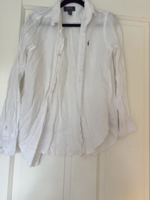 Vit linneskjorta från Polo Ralph Lauren - Klassisk vit skjorta i linne från Polo Ralph Lauren med knappar framtill och lång ärm. Skjortan har en liten broderad logga på bröstet och en avslappnad passform. Perfekt för en stilren och fräsch look.
