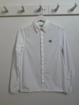Vit skjorta från Kenzo med tigerlogo - Klassisk vit skjorta från Kenzo med lång ärm och snygg broderad tigerlogo på bröstet. Skjortan har knappar framtill och är gjord i bomull för en clean och stilren look. Perfekt till jeans eller kostymbyxor. Nypris 2600.