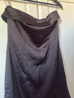 Svart axelbandslös satinklänning - Snygg svart axelbandslös klänning i glansigt satintyg med draperad urringning och slits nedtill. Klänningen har en elegant och enkel design som ger en lyxig känsla. Perfekt för dig som vill sticka ut med en stilren look. Endats använt en gång innan så i väldigt fint skick!! passar jätte bra till sormiga dagar men även till mer speciella event !! (pris går självklart att justeras)