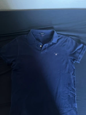 Mörkblå pikétröja från GANT - Klassisk mörkblå pikétröja från GANT i bomull med krage och knappar framtill. Tröjan har en liten broderad logga på bröstet och en slim passform som ger en clean och stilren look.
