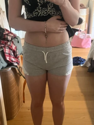 Grå shorts med spetsdetaljer - Grå shorts med vit snörning i midjan och fina spetsdetaljer på sidorna. Mjukt material och elastisk midja för skön passform. Perfekta för varma dagar eller som mysplagg hemma. Använda mycket men inte synliga skador. 