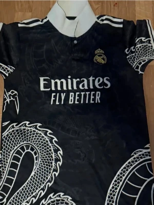 Real Madrid svart matchtröja drakmönster - Snygg svart Real Madrid matchtröja från Adidas med vitt drakmönster och klubbmärke på bröstet. Tröjan har vit krage, korta ärmar och vita detaljer på axlarna. Emirates Fly Better-logga tryckt på framsidan. Perfekt för fotbollsfans!