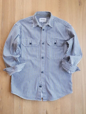 Randig overshirt från Carhartt - Säljer en blåvit randig overshirt från Carhartt med två bröstfickor och knappar framtill. Overshirten har klassisk krage, långa ärmar och är gjord i slitstark bomull. Perfekt lager-på-lager-plagg för en avslappnad stil.
