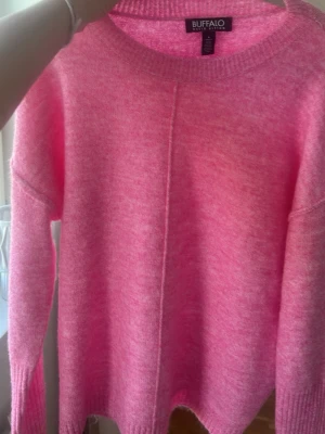 Rosa stickad tröja Buffalo - Säljer en fluffig rosa stickad tröja från Buffalo David Bitton. Tröjan har rund halsringning, lång ärm och en dekorativ söm mitt fram. Perfekt för dig som gillar färg och vill ha något mjukt och bekvämt i garderoben.