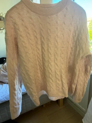 Ljusrosa kabelstickad tröja - Mysig ljusrosa kabelstickad tröja med rund halsringning och långa ärmar. Tröjan har ett klassiskt flätmönster och ribbade muddar vid ärmslut och nederkant. Perfekt för att hålla värmen under kyliga dagar.