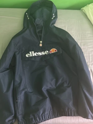 Mörkblå vindjacka från Ellesse - Snygg mörkblå vindjacka från Ellesse med stor logga på bröstet. Jackan har huva med dragsko, halv dragkedja framtill och en stor magficka. Tillverkad i ett lätt och vattenavvisande material, perfekt för blåsiga dagar. Pris kan sänkas säljer den minst för 400kr !!!