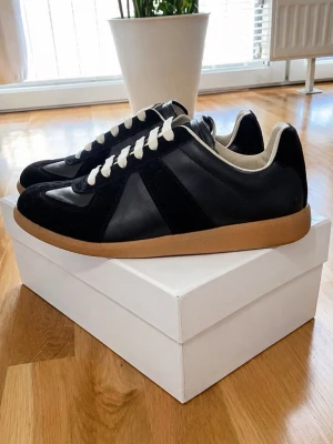 Maison Margiela Gats - Stilrena svarta sneakers från Maison Margiela med beige sula och vita snören. Ovandelen är i en mix av mocka och läder, vilket ger en lyxig känsla. Klassisk low-top siluett som passar till många olika outfits.