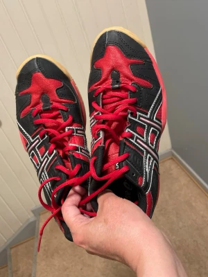 Röda och svarta Asics inomhusskor - Snygga inomhusskor från Asics i rött och svart med vita detaljer och röda skosnören. Skorna har en klassisk sportig design med slitstark ovandel i mesh och syntetmaterial samt greppvänlig gummisula. Perfekta för träning eller sporthall.