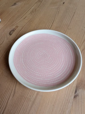 Marimekko oiva tallrik med rosa Räsymatto - Säljer en rund tallrik i keramik med vitt yttre och ett ljusrosa cirkulärt mönster på insidan. Perfekt för att piffa upp köket eller servera fika på ett snyggt sätt. Tallriken har en modern och stilren design. 