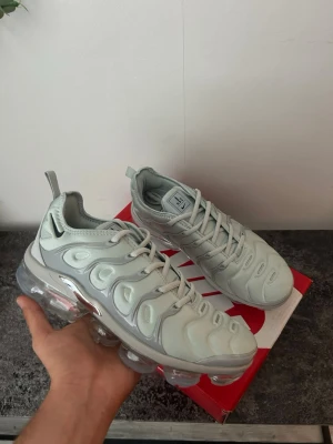 Nike Air Vapormax Plus ljusgrå - Nike Air Vapormax Plus sneakers i ljusgrått med en futuristisk design och genomskinlig sula. Ovandelen är i syntetmaterial med plastdetaljer och tydliga linjer. Swoosh-logga på hälen och dragflik bak. Perfekta för dig som gillar streetwear och sportig stil.