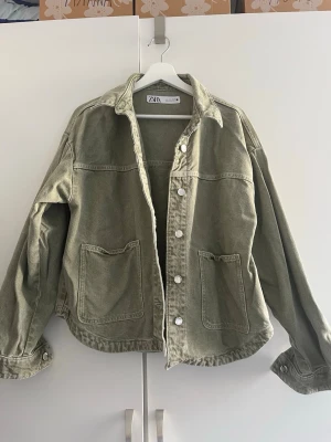 Olivgrön jeansjacka från Zara - Snygg olivgrön jeansjacka från Zara med avslappnad passform. Jackan har två stora framfickor, klassisk krage och silverfärgade knappar. Perfekt för dig som gillar en cool och enkel stil. Materialet är robust jeans som ger en trendig look.