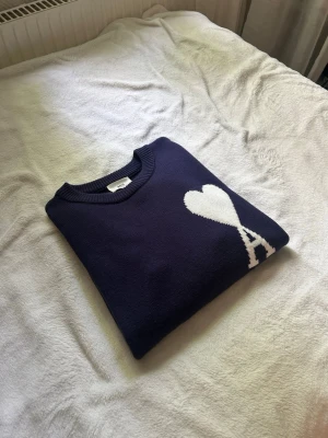 Mörkblå stickad tröja från Ami Paris - Snygg mörkblå stickad tröja från Ami Paris med ett vitt hjärta och bokstaven A på bröstet. Tröjan har rund halsringning och ribbade muddar. Perfekt för dig som gillar stilrena detaljer och vill ha något som sticker ut lite extra.