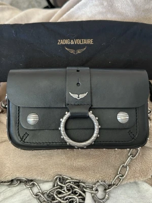 Zadig & Voltaire svart handväska - Cool svart handväska i läder från Zadig & Voltaire med silverfärgade metalldetaljer, stor ring framtill och kedjerem. Väskan har ett emblem med vingar och dekorativa nitar på sidorna. Perfekt accessoar för dig som gillar edgy och unik design. Original pris är 6 729kr !! 💕