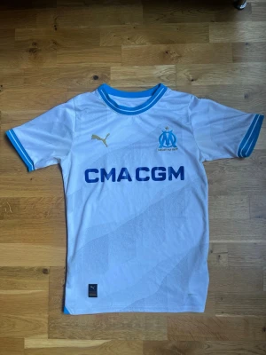 Marseille matchtröja Puma vit/blå - Säljer en vit Olympique de Marseille matchtröja från Puma med blå detaljer och klubbmärke på bröstet. Tröjan har korta ärmar, rund hals och tryckt sponsorlogga framtill. Perfekt för dig som älskar fotboll och vill stötta ditt lag.