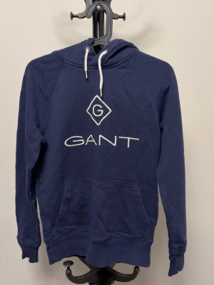 Blå hoodie från GANT med logga - Mörkblå hoodie från GANT med stor vit broderad logga och emblem på bröstet. Klassisk känguruficka framtill och vit dragsko i huvan. Tillverkad i mjukt bomullsmaterial och har ribbade muddar vid ärmslut och nederkant.