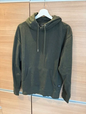 Olivgrön hoodie från Jack & Jones - Säljer en olivgrön hoodie från Jack & Jones med klassisk känguruficka och justerbar huva med dragsko. Hoodien är tillverkad i mjuk bomullsblandning och har ribbade muddar vid ärmslut och nederkant. Perfekt för en avslappnad och stilren look.