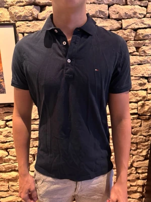 Marinblå pikétröja från Tommy Hilfiger - Snygg marinblå pikétröja från Tommy Hilfiger med klassisk krage och knappar framtill. Liten logga på bröstet och korta ärmar. Tillverkad i mjuk bomull för en bekväm känsla och stilren look.