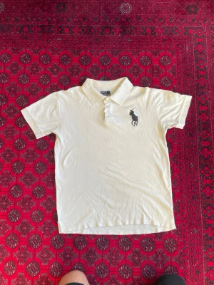 Vit pikétröja från Ralph Lauren - Klassisk vit pikétröja från Ralph Lauren med korta ärmar och krage. Tröjan har två knappar vid halsen och den ikoniska mörkblå broderade loggan på bröstet. Perfekt till jeans eller shorts för en clean och stilren look.