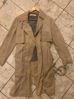 Beige trenchcoat med bälte från Zara  - Stilren beige trenchcoat med klassisk krage och avtagbart bälte i midjan. Jackan har rutigt foder i svartvitt och långa ärmar. Perfekt för dig som gillar tidlös och enkel stil. Materialet ser ut att vara bomull eller en bomullsblandning. Är mycket bra skick! 