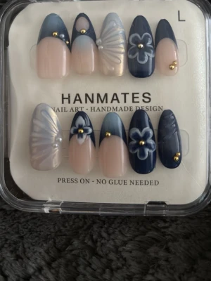 Blå och nude press-on naglar HANMATES - Snygga press-on naglar i blått och nude från HANMATES. Naglarna har mandelform och dekoreras med blommor, pärlor och guldiga detaljer. Designen är handgjord och materialet är plast. Ingen lim behövs, superenkelt att applicera!