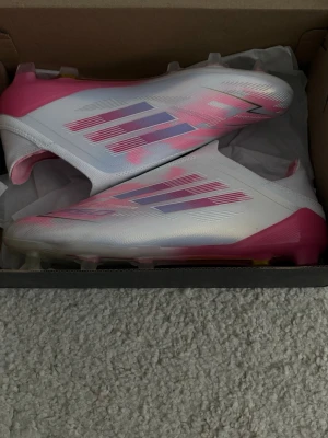 Adidas vita och rosa elites F50s - Säljer ett par helt nya grymma fotbollsskor från Adidas i vitt och rosa med metallic finish och ikoniska tre ränder på sidan. Skorna har en strömlinjeformad design utan snörning och är gjorda i syntetmaterial för bästa grepp och komfort på planen.