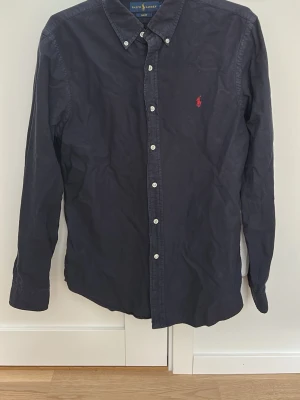 Mörkblå skjorta från Ralph Lauren - Klassisk mörkblå skjorta från Ralph Lauren med vit knappstängning och röd broderad logga på bröstet. Skjortan har lång ärm och button-down krage. Perfekt till jeans eller chinos för en clean och stilren look.