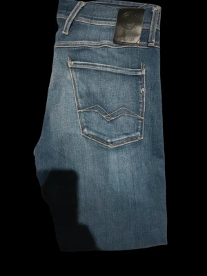 Replay ”Anbass” 34/32 - Snygga blå jeans från Replay med raka ben och klassisk femficksmodell. Jeansen har ljus tvätt och kontrastsömmar, samt en unik detalj på bakfickan. Perfekta för en avslappnad och trendig stil.